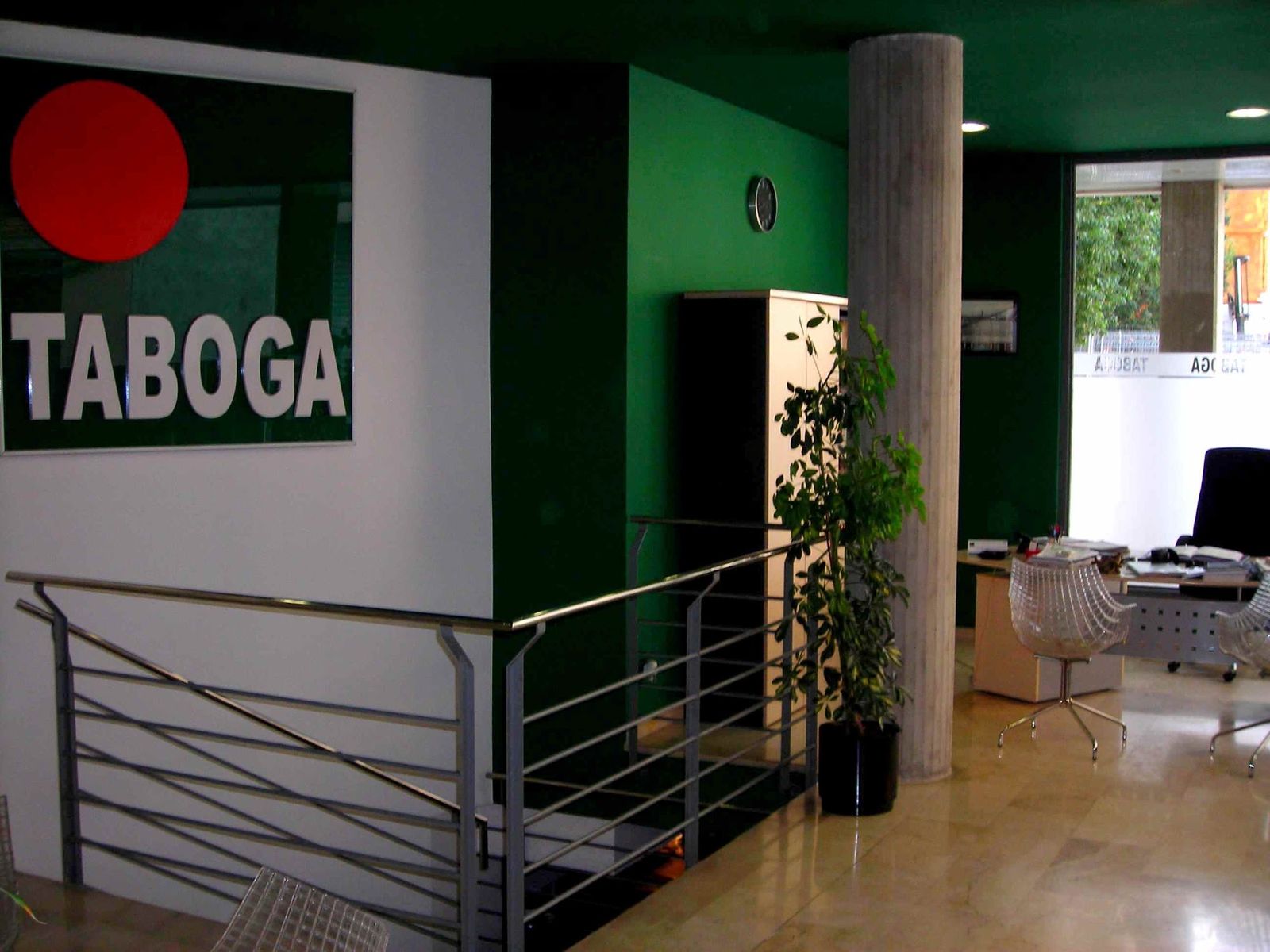 Interior de la oficina: paredes verdes y blancas, el cartel TABOGA, la barandilla de acero y las mesas